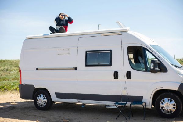 Biały kamper w podróży – marzenie miłośników caravaningu Turystka z lornetką siedzi na dachu białego kampera zaparkowanego podczas podróży